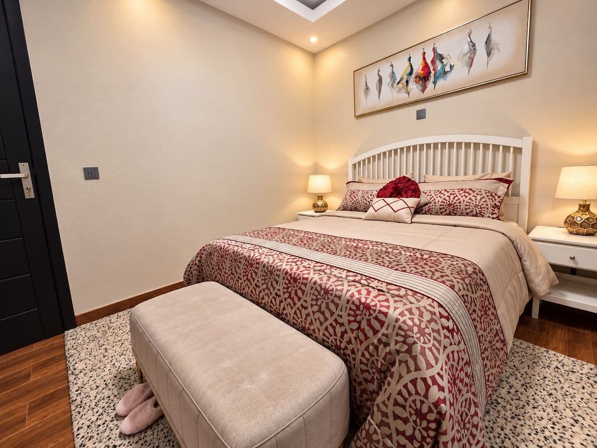 A bedroom with a white slatted headboard and red-patterned bedding. Irebero, Rebero.