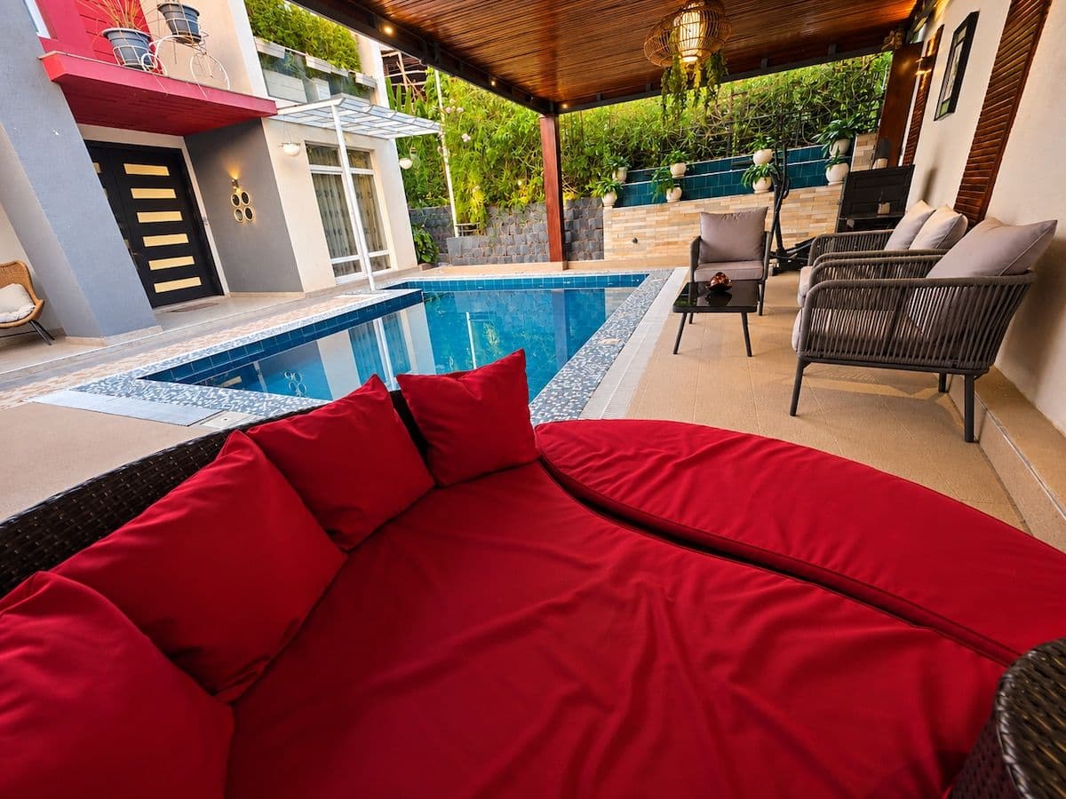 A red daybed with cushions overlooking the pool and the covered lounge. Irebero, Rebero.